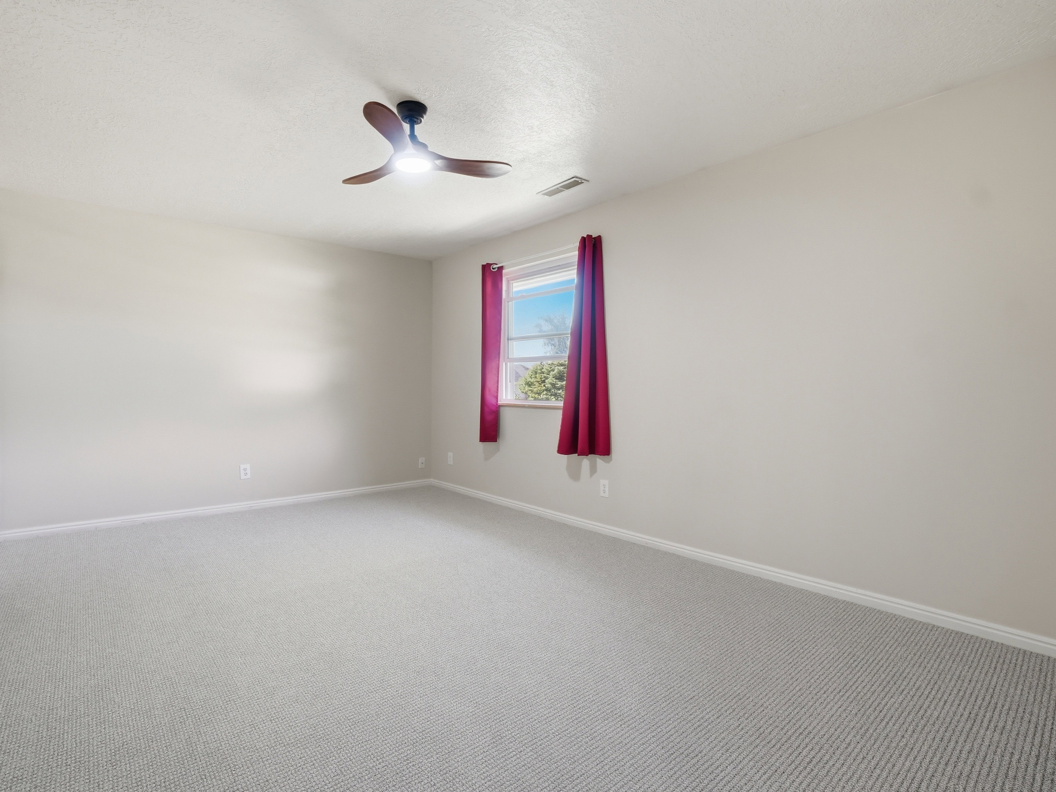 Spacious room featuring light-toned carpeting, neutral wall finishes, a ceiling fan with integrated lighting, a single window, and baseboard heating