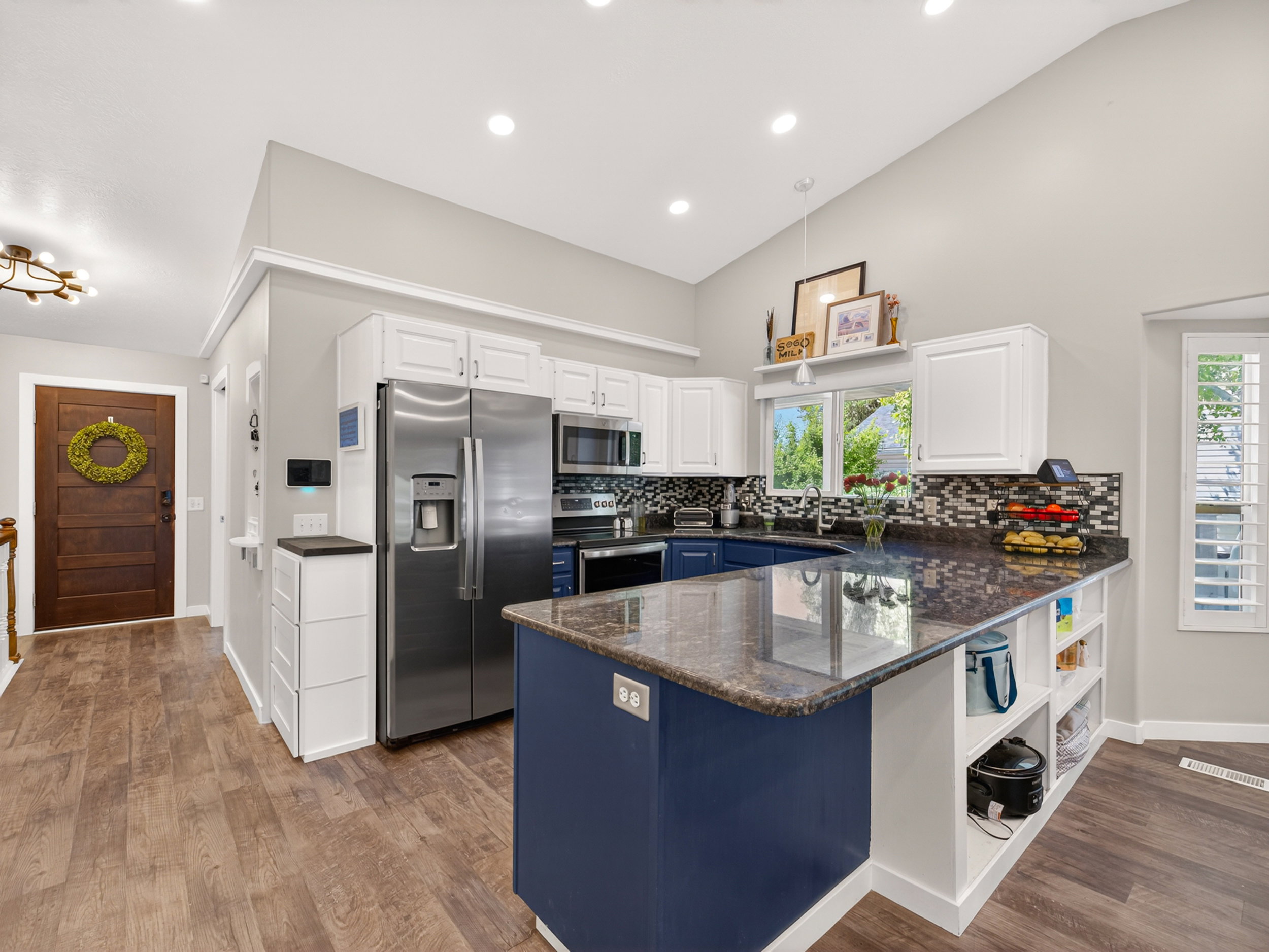 Contemporary kitchen featuring a vaulted ceiling, wood-finish flooring, white upper cabinetry, a blue island with built-in shelving, and stainless steel appliances