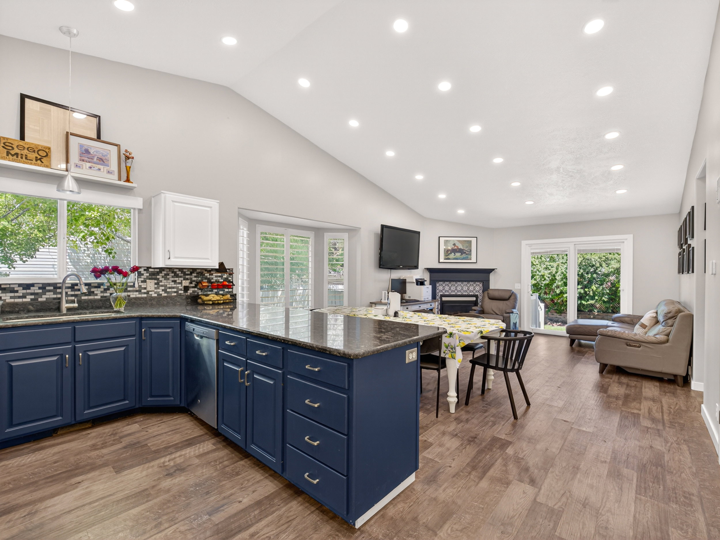 Open concept living space featuring a vaulted ceiling with recessed lighting, wood-finish flooring, and a kitchen with blue cabinetry, dark countertops, and a mosaic tile backsplash