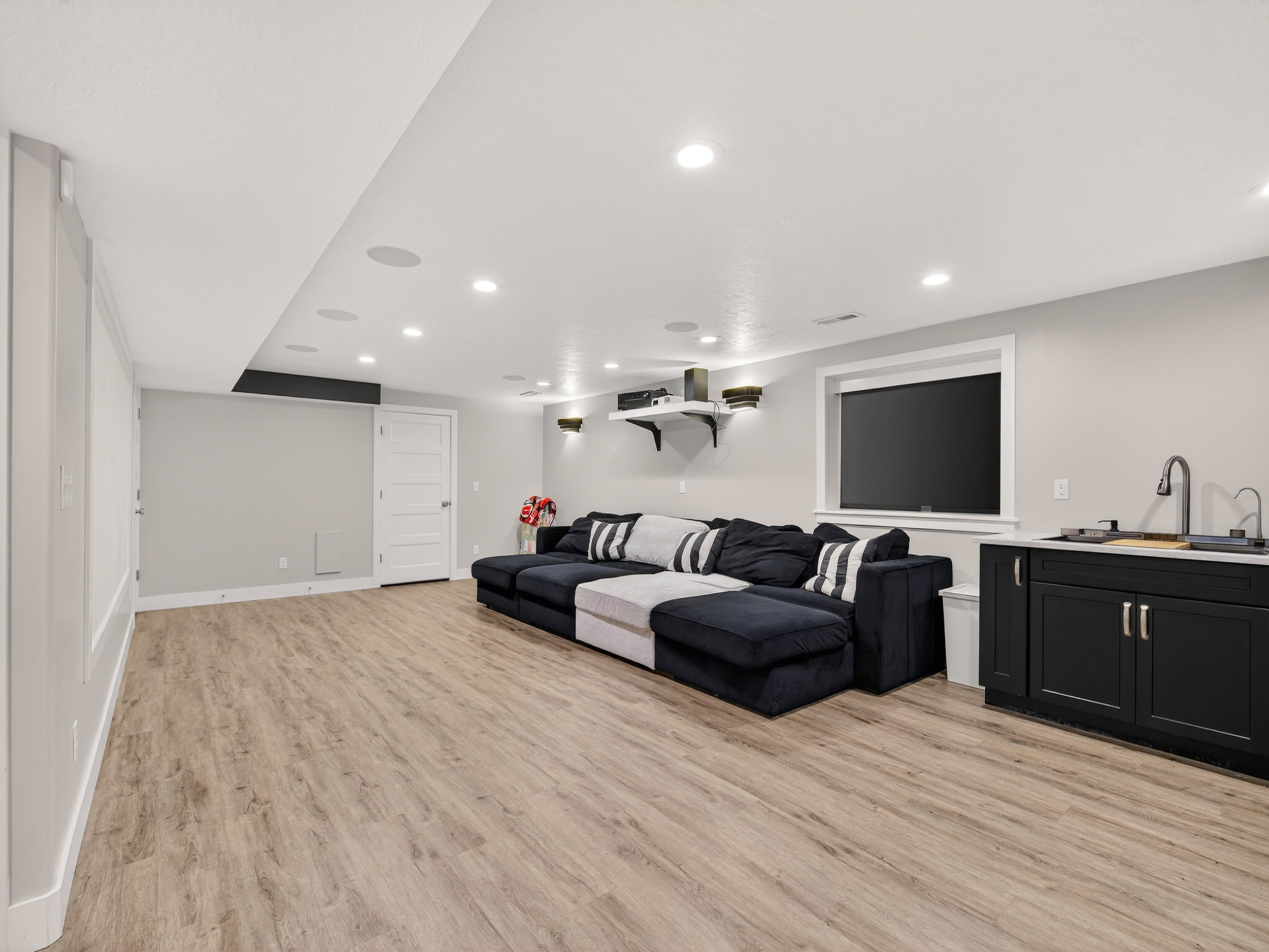 Spacious entertainment area featuring wood-finish flooring, recessed lighting, and a built-in kitchenette with a dark cabinetry and a white countertop