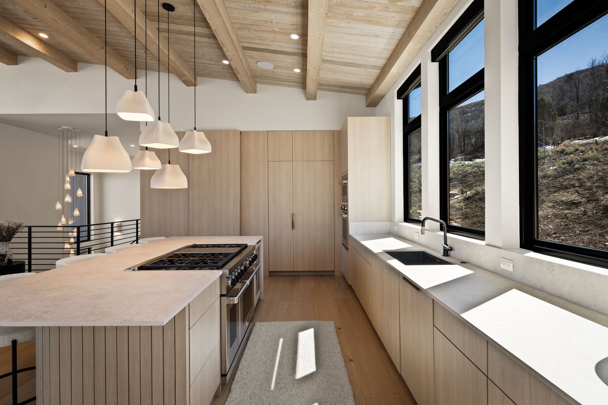Kitchen featuring an expansive center island with an integrated gas range, light wood-finish cabinetry, a deep basin sink with a gooseneck faucet, light-toned countertops, and multiple large windows with black frames