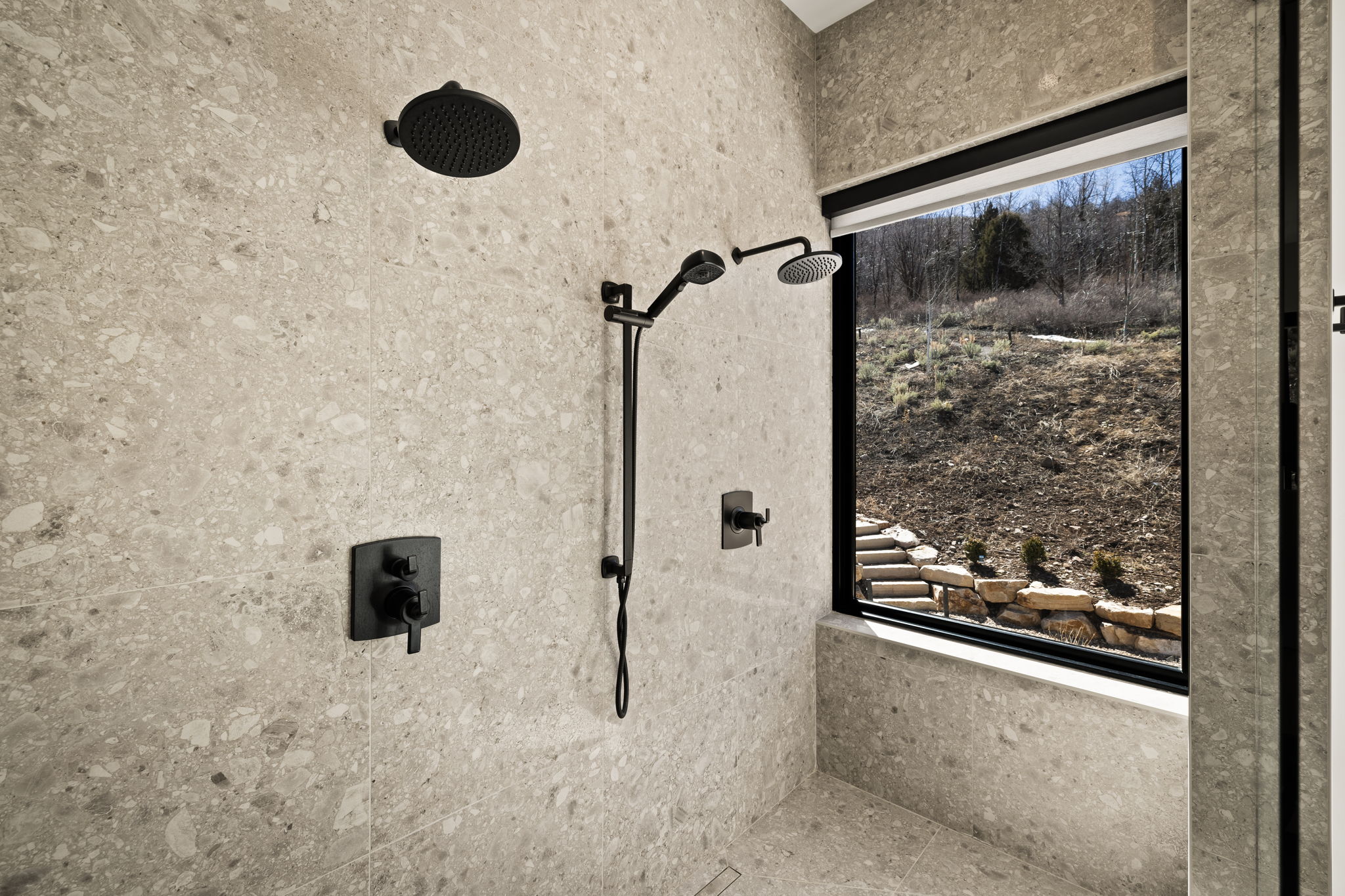 Walk-in shower with full-height stone-finish tiling, matte black fixtures including a rain shower head and handheld shower, and a large window