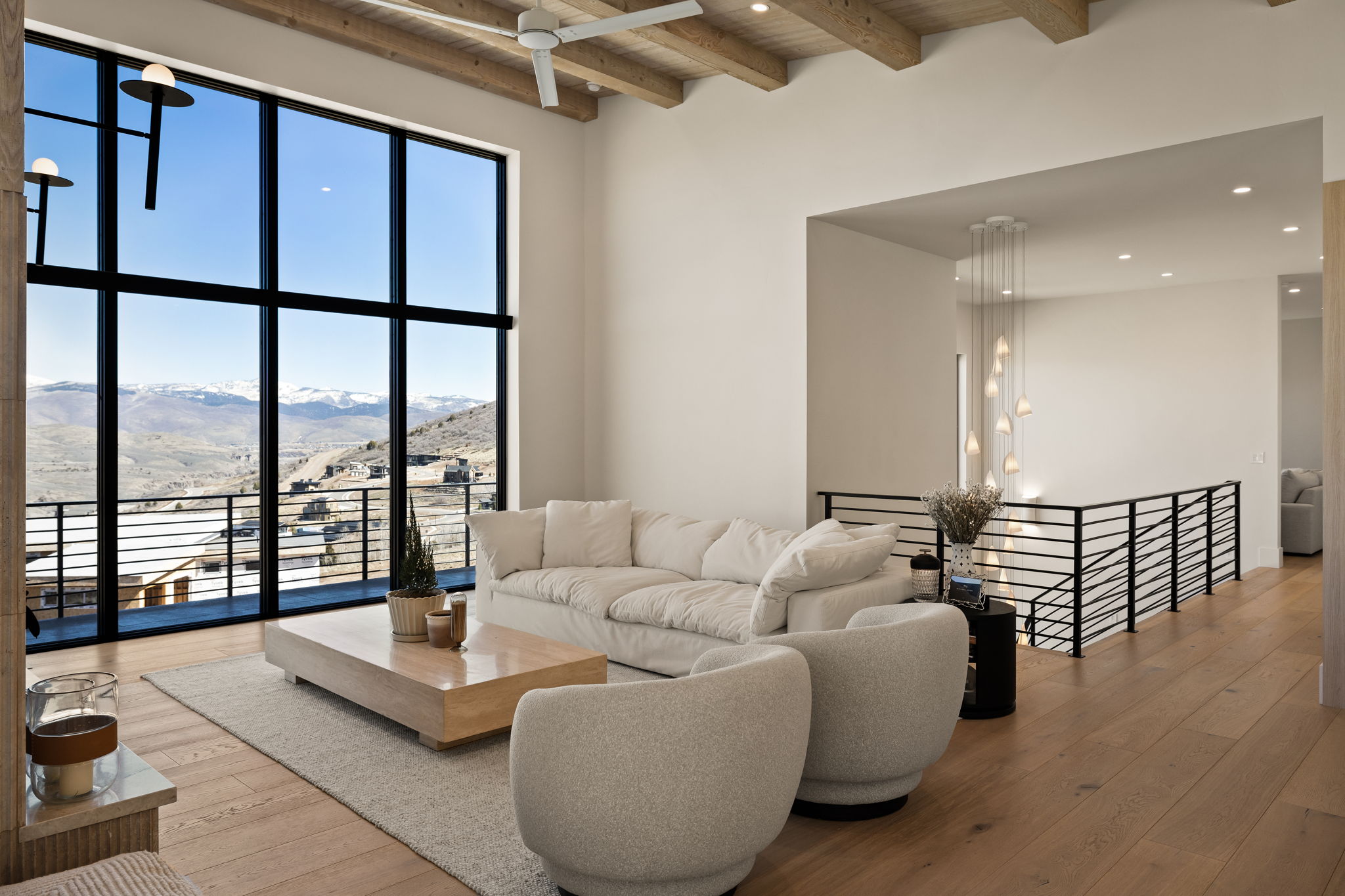 Open-concept living area featuring a vaulted ceiling with exposed wood beams, expansive floor-to-ceiling windows, light wood-finish flooring, and a contemporary multi-pendant light fixture over the staircase