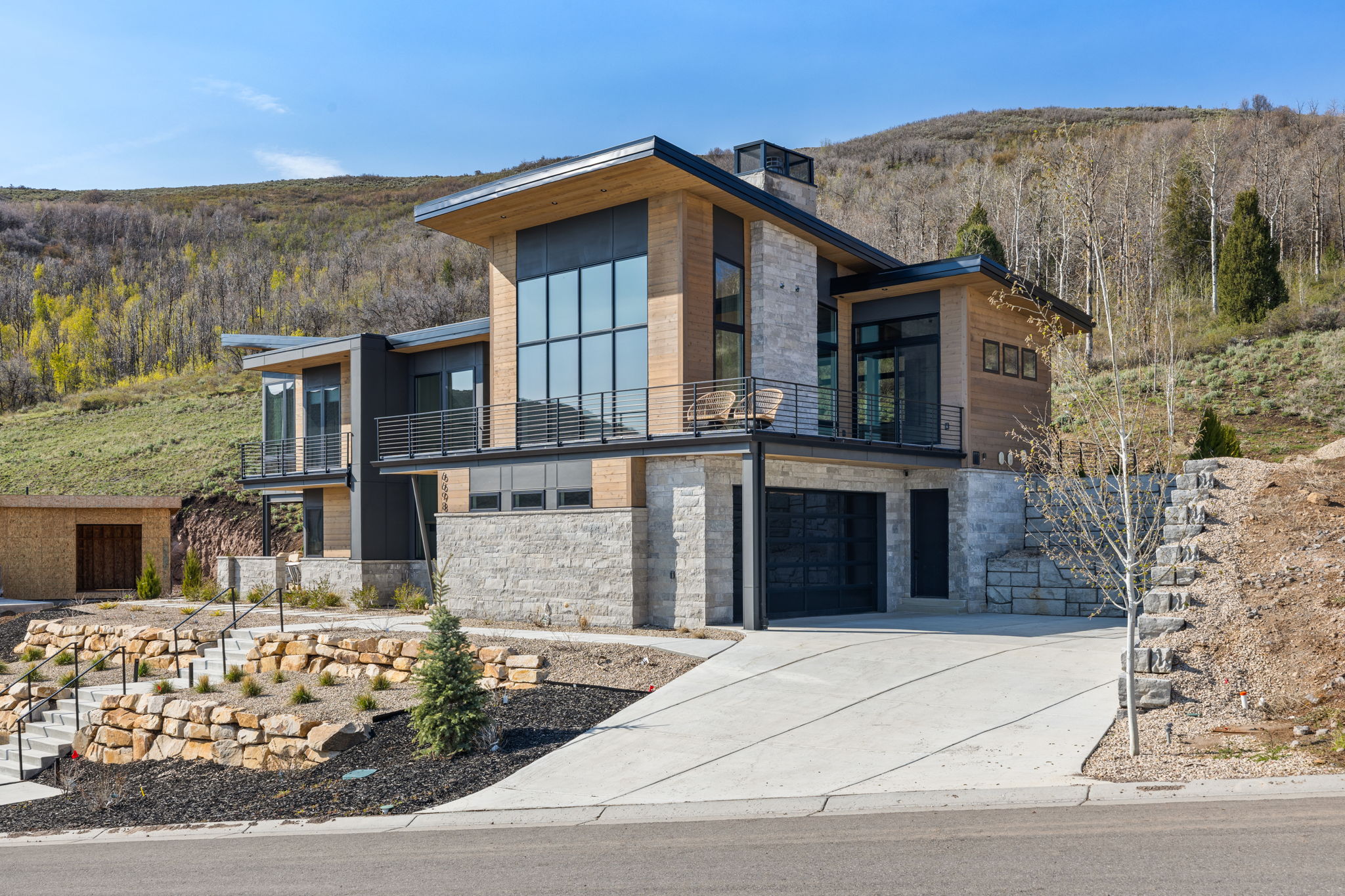Modern architectural design featuring extensive window walls, stone facade accents, wood-finish paneling, multiple balconies with metal railings, and an integrated garage with a dark door