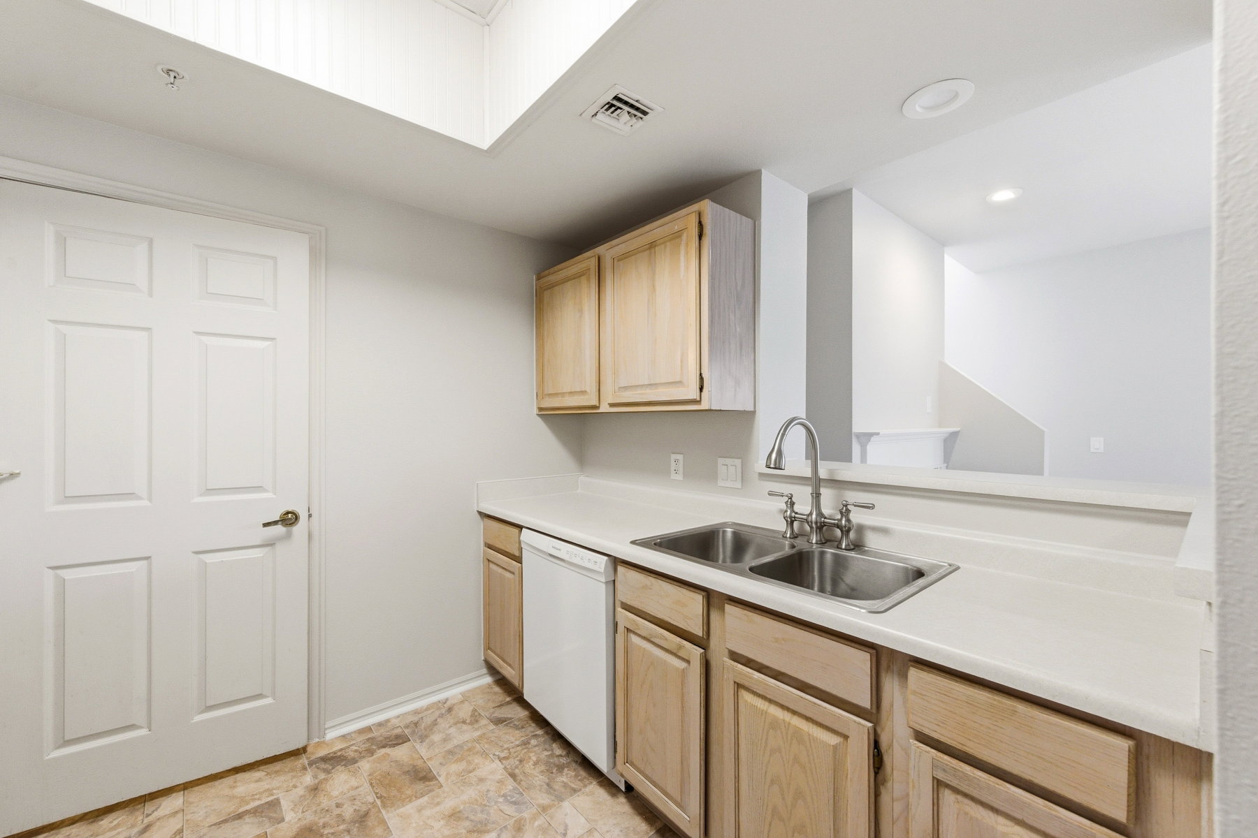Kitchen area featuring light wood-finish cabinetry, a double basin stainless steel sink with a gooseneck faucet, a white dishwasher, light-toned countertops, and patterned floor tiling