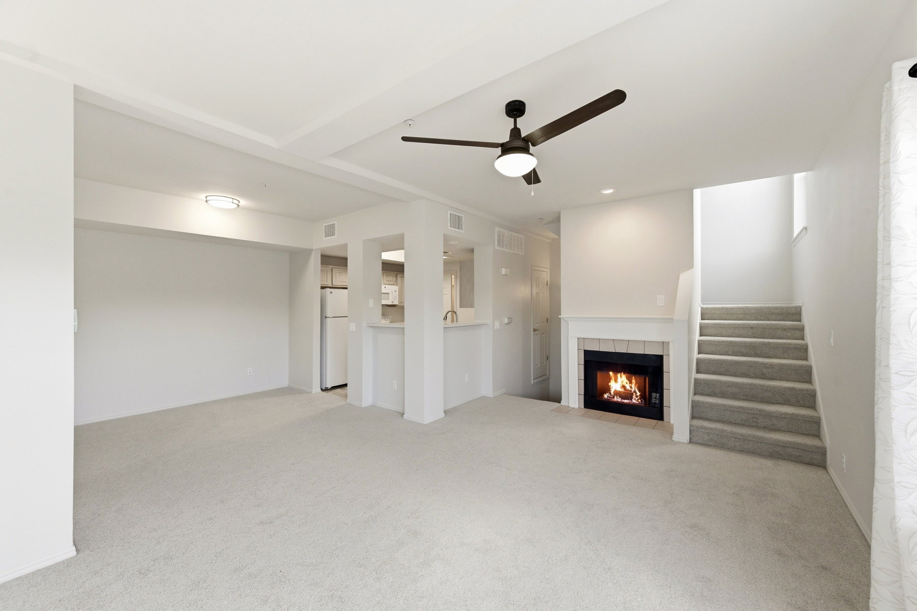 Carpeted living area featuring a fireplace with a white mantel, ceiling fan, and a light-toned kitchen with white appliances