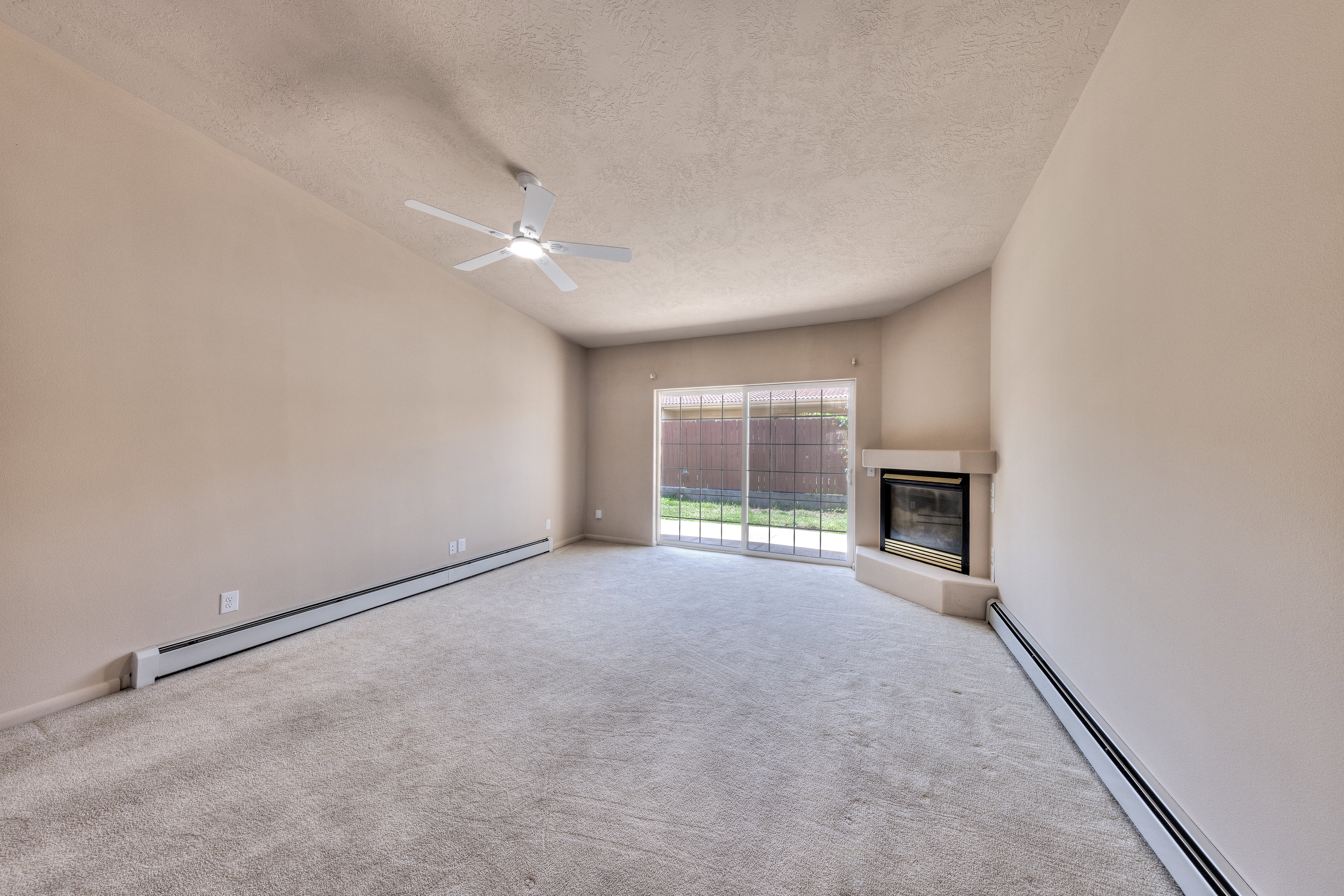 Carpeted living area featuring a corner fireplace with a light-toned surround, a ceiling fan, and sliding glass doors opening to a fenced outdoor area with grass