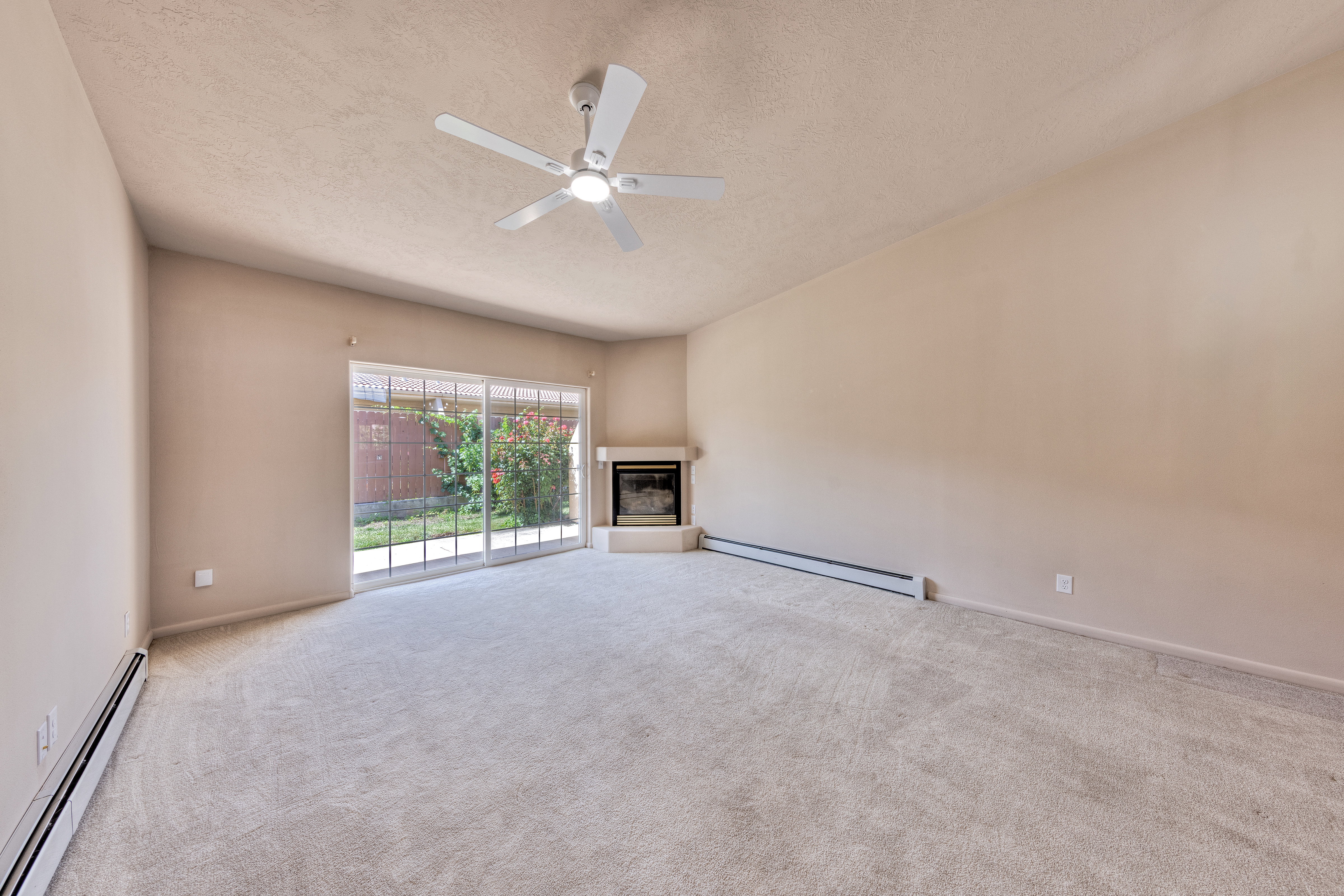 Spacious carpeted room featuring a ceiling fan, a corner fireplace with a hearth, and a sliding glass door leading to a fenced outdoor area
