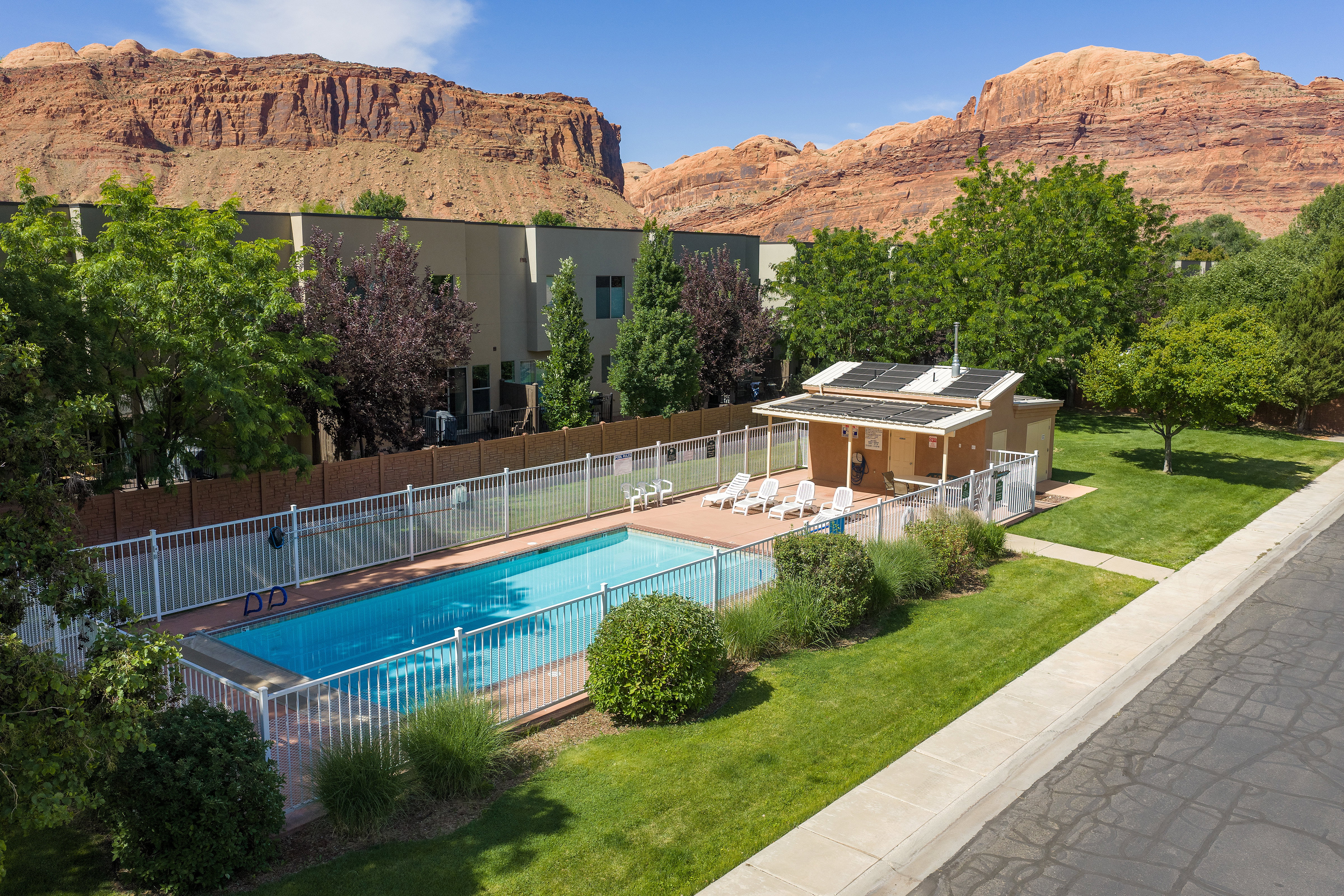 Fenced swimming pool area featuring a pool house with solar panels, surrounded by a manicured lawn and mature trees