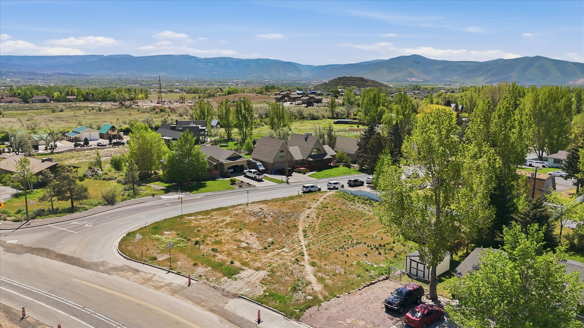 Expansive undeveloped lot presenting a prominent corner location with paved road frontage, mature trees, and mountainous terrain in the distant background
