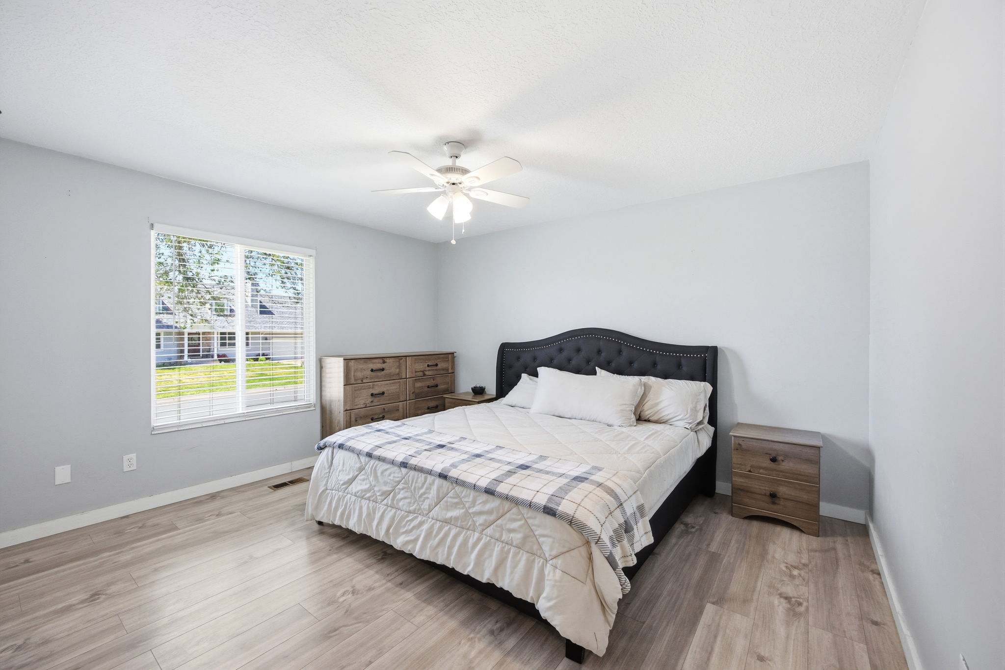 Bright bedroom featuring light wood-finish flooring, a large window with blinds, a ceiling fan with light fixture, and light gray wall paint