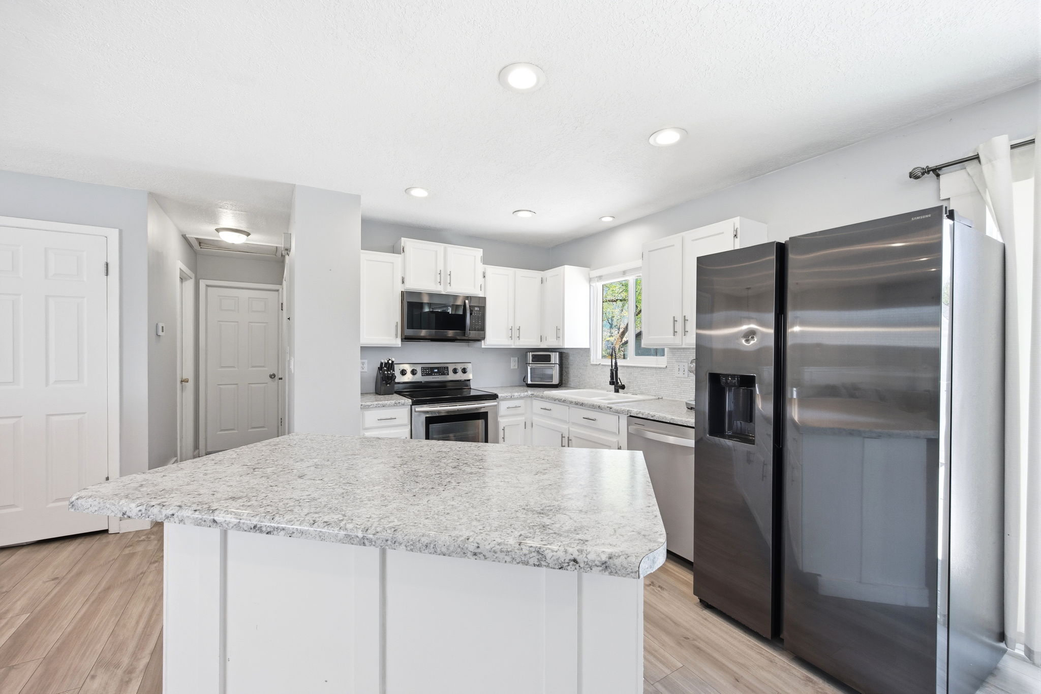 Kitchen featuring a central island with a light-toned countertop, white cabinetry, integrated stainless steel appliances, recessed lighting, and wood-finish flooring