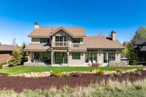 Rear exterior featuring extensive glazing, board-and-batten siding, multiple patios, a second-story balcony, and a stone-clad chimney