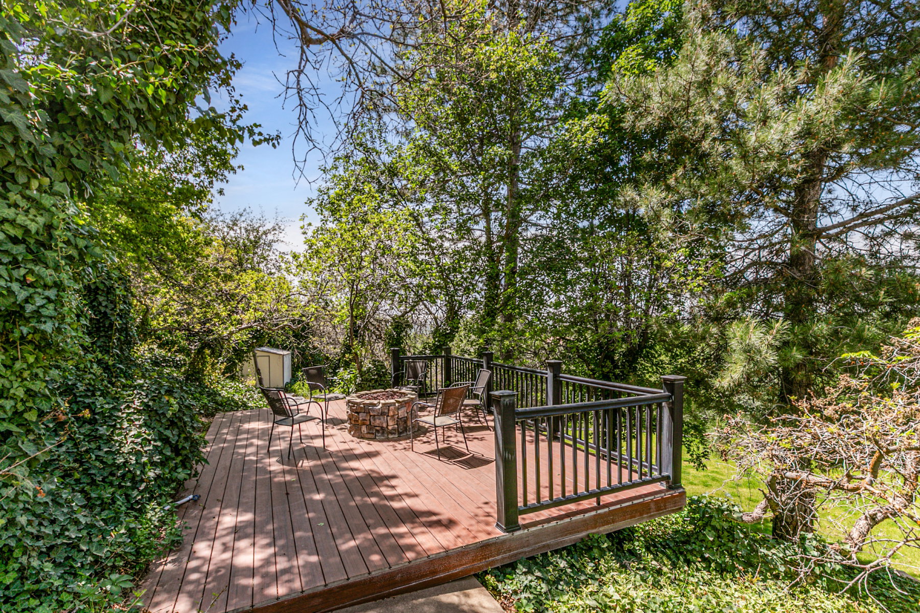Expansive wood-finish deck featuring a stone-clad fire pit, black metal railings, and lush surrounding foliage