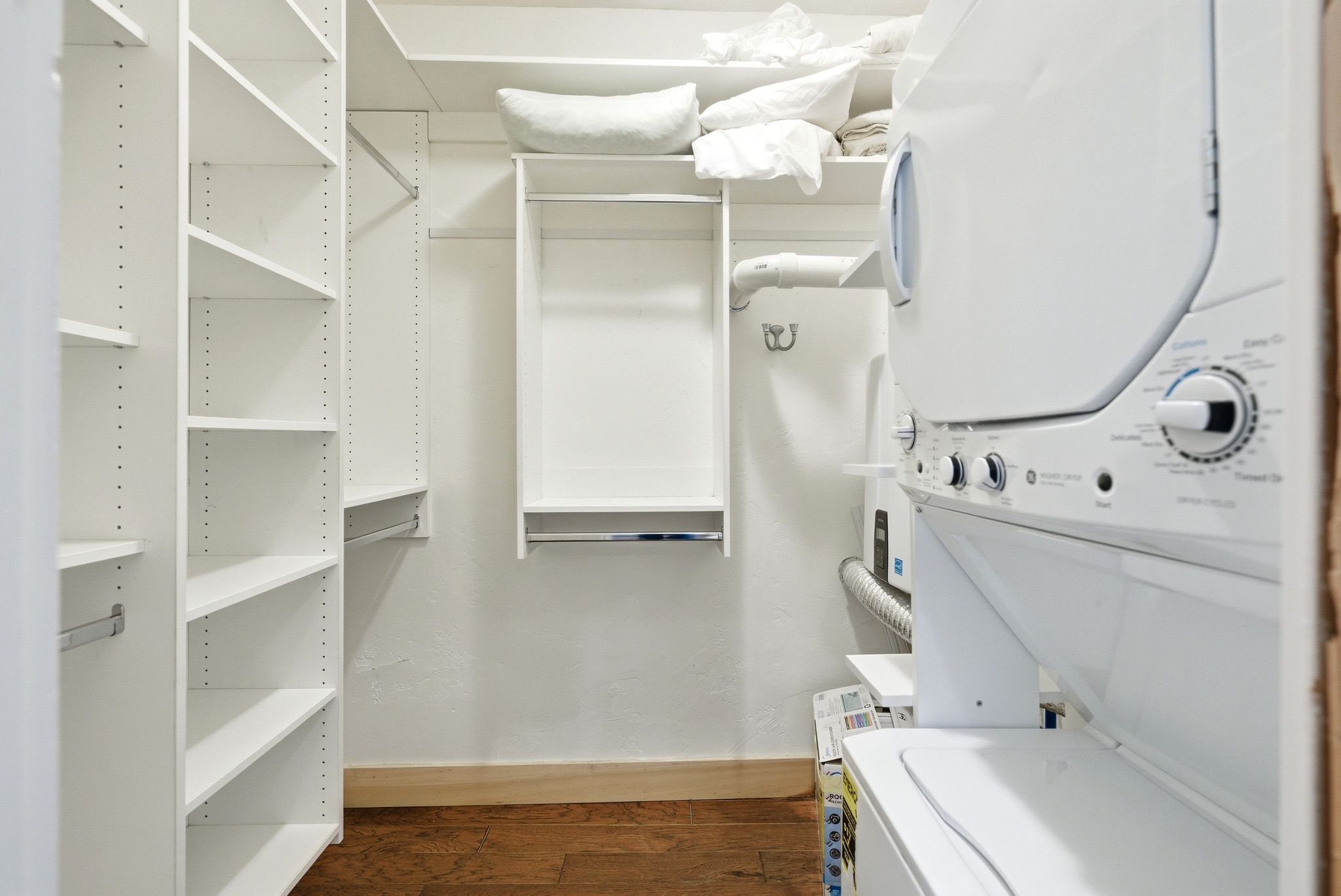 Integrated laundry closet featuring a stacked washer and dryer, white adjustable shelving, and wood-finish flooring