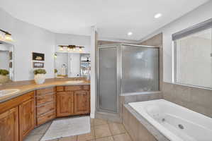 Bathroom featuring a jetted soaking tub, glass-enclosed shower, dual vanities with integrated sinks, wood-finish cabinetry, and neutral tile flooring