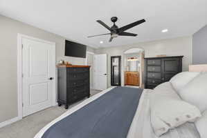 Carpeted bedroom featuring a contemporary ceiling fan with integrated lighting, recessed ceiling lights, an arched doorway, and a bathroom entry with a wood-finish vanity and mirror