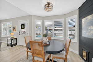 Dining area featuring a contemporary round table, light wood-finish flooring, a modern orb chandelier, multiple windows with white plantation shutters, and a dark accent wall with a built-in fireplace