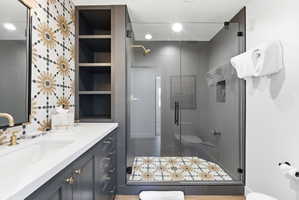 Bathroom featuring a glass-enclosed shower with decorative floor tiling, a vanity with dual sinks, a quartz countertop, and gold-finish fixtures