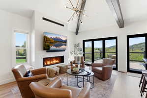 Living area featuring a modern linear fireplace, high vaulted ceilings with exposed beams, wood-finish flooring, and multiple glass doors opening to an outdoor deck