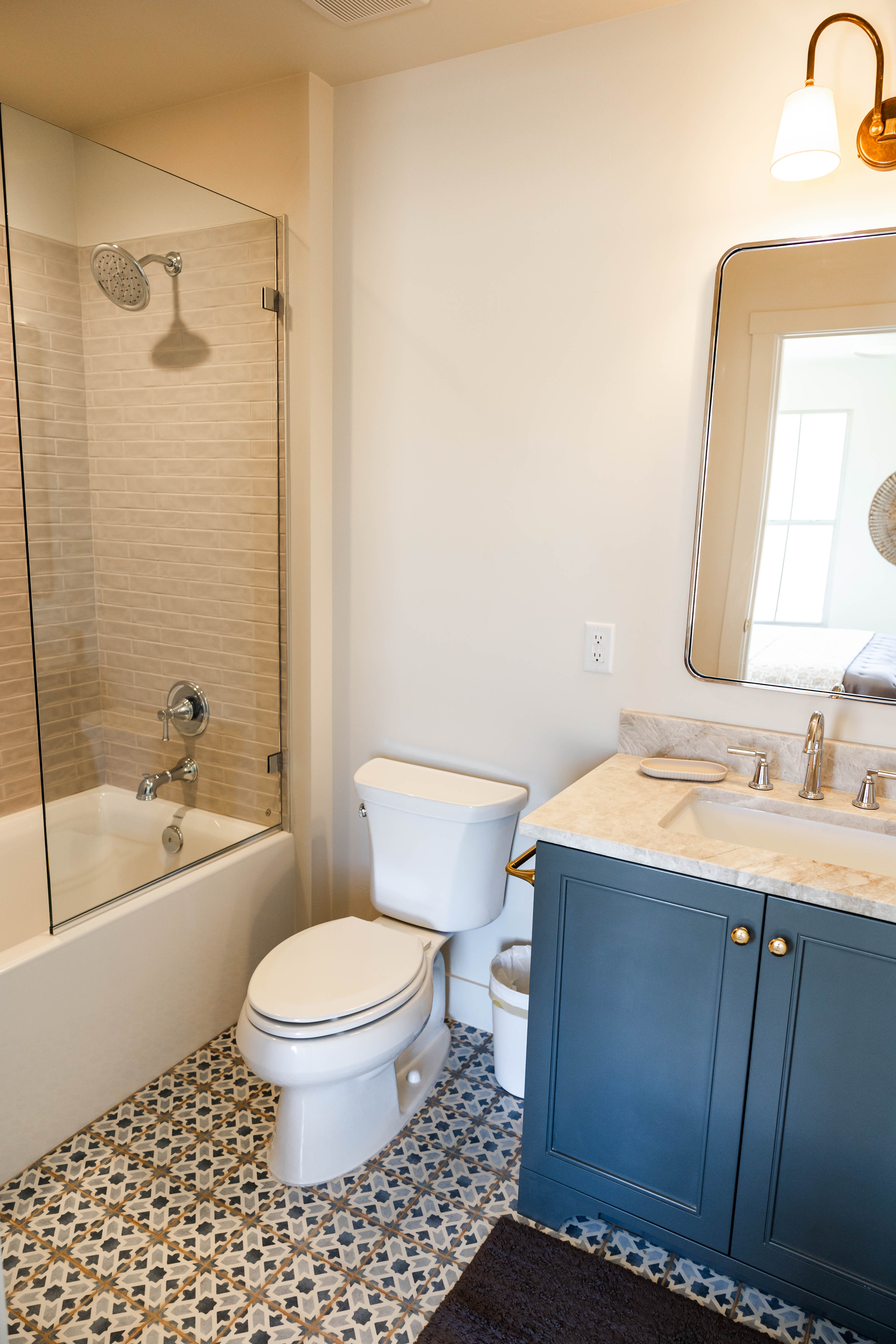 Bathroom featuring patterned floor tiling, a glass-enclosed tub/shower combo with subway tile surround, a dark blue vanity with gold hardware, a stone countertop, and a wall-mounted mirror with an overhead light fixture
