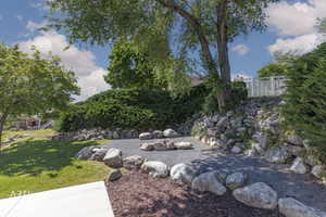 Outdoor gravel seating area with natural rock accents, surrounded by lush green landscaping, mature trees, and a retaining wall