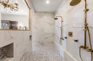 Spacious shower featuring white marble-look wall tiles, pebble-style floor tiling, and dual brass-finish shower systems
