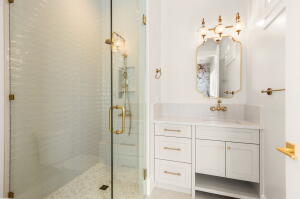 Bathroom featuring a clear glass shower enclosure with brass-finish hardware, a white vanity with drawers and cabinet storage, a stone-finish countertop, and a decorative wall-mounted mirror with an ornate light fixture