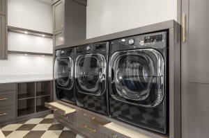 Dedicated laundry space featuring three front-load appliances, checkerboard flooring, built-in cabinetry, open shelving with integrated lighting, and countertop space