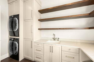 Laundry area featuring stacked appliances, cream cabinetry, light-toned countertop with integrated sink, and wood-finish open shelving