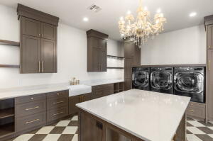 Dedicated laundry room featuring an overhead crystal chandelier, checkerboard flooring, a deep basin sink, extensive cabinetry, and a central island with a solid surface countertop