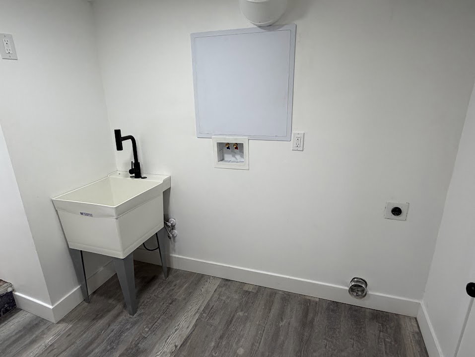 Dedicated laundry space featuring a utility sink with a matte black faucet, wall-mounted electrical outlets, and wood-finish flooring
