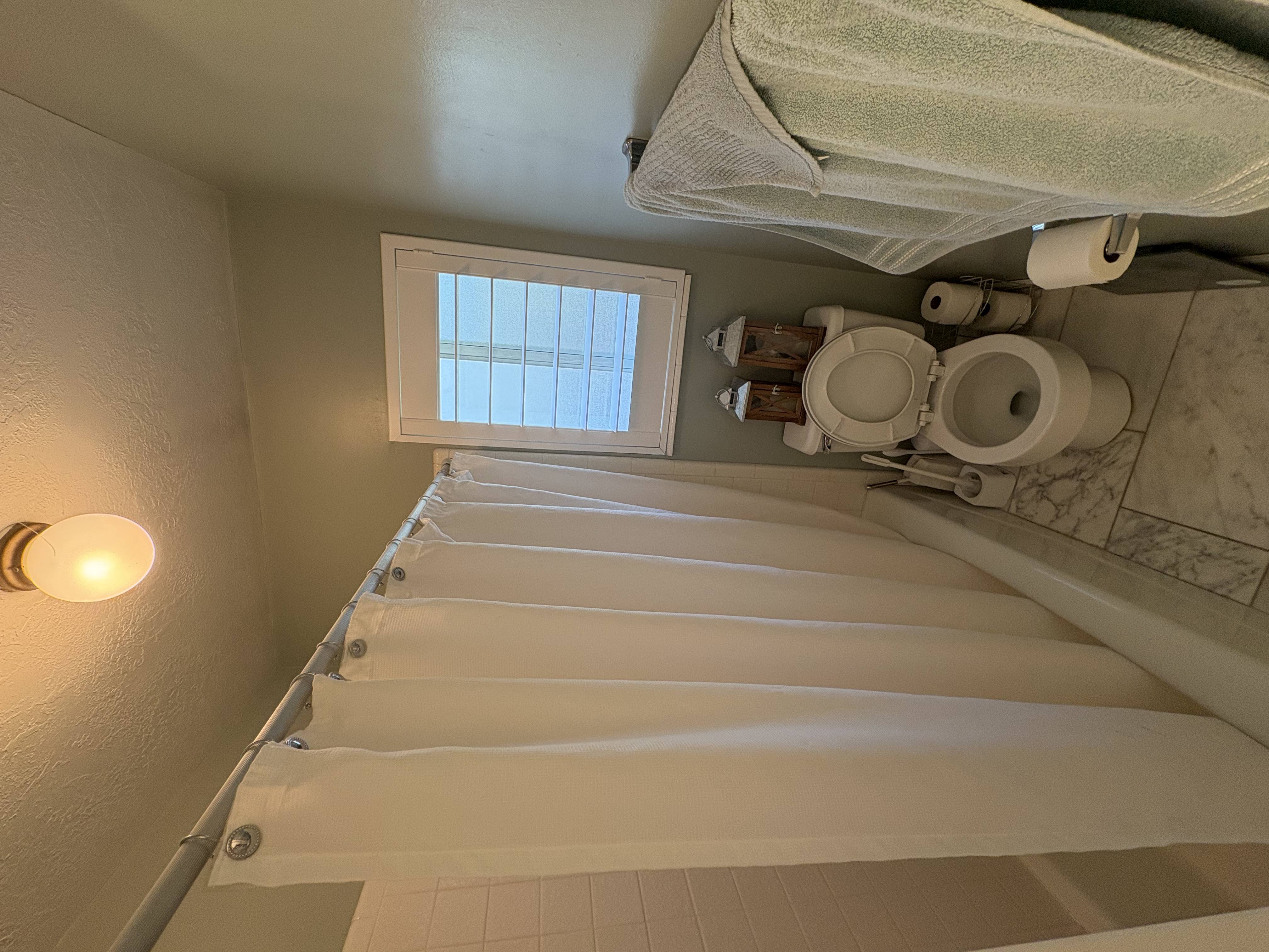 Bathroom featuring a full tub with shower, white tile surround, window with white plantation shutters, and a toilet with white marble-look floor tiling