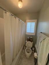 Bathroom featuring a full tub with shower, white tile surround, window with white plantation shutters, and a toilet with white marble-look floor tiling