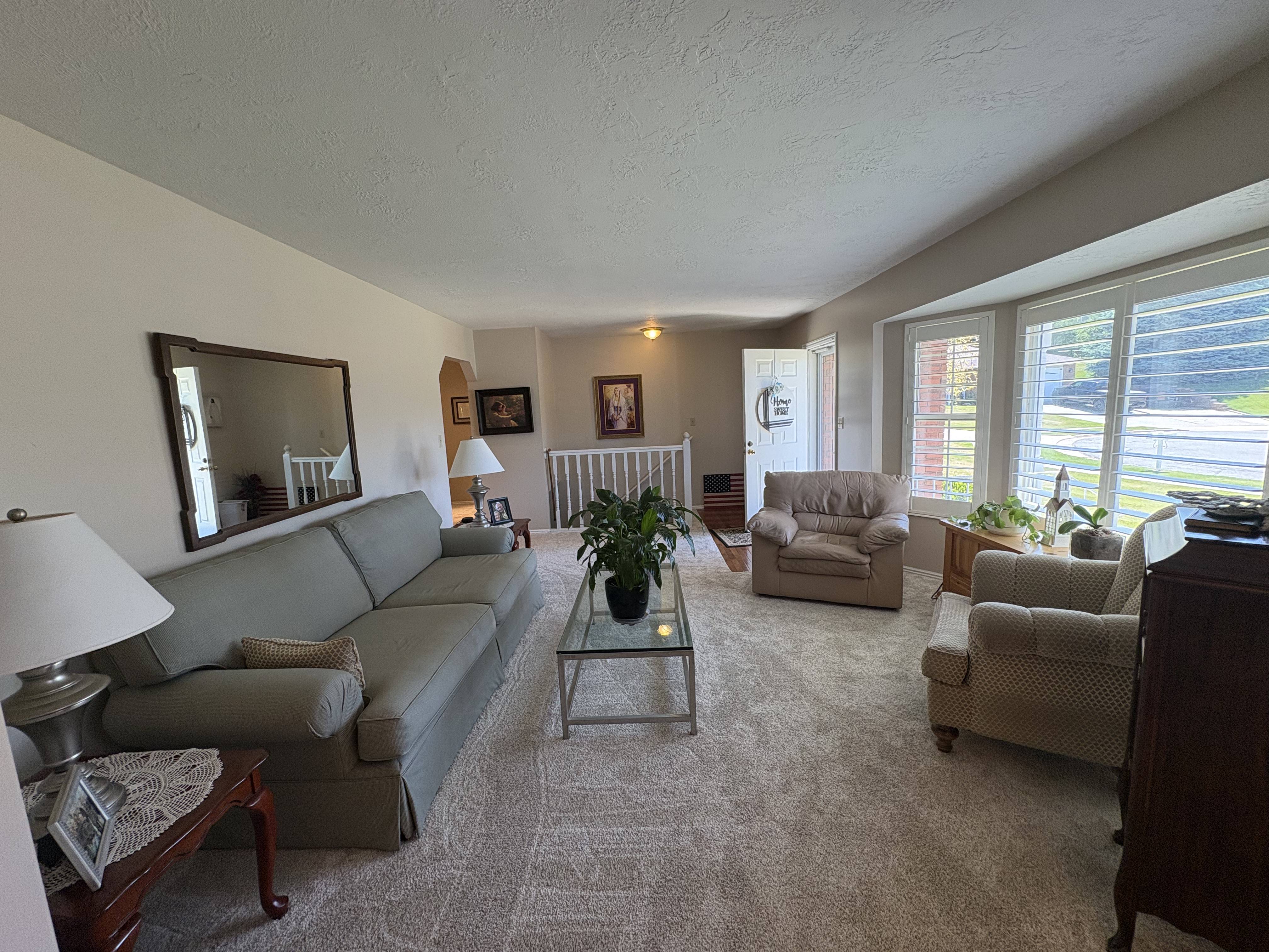 Living area featuring neutral wall tones, textured carpeting, a bay window with plantation shutters, a glass-top coffee table, and an arched doorway