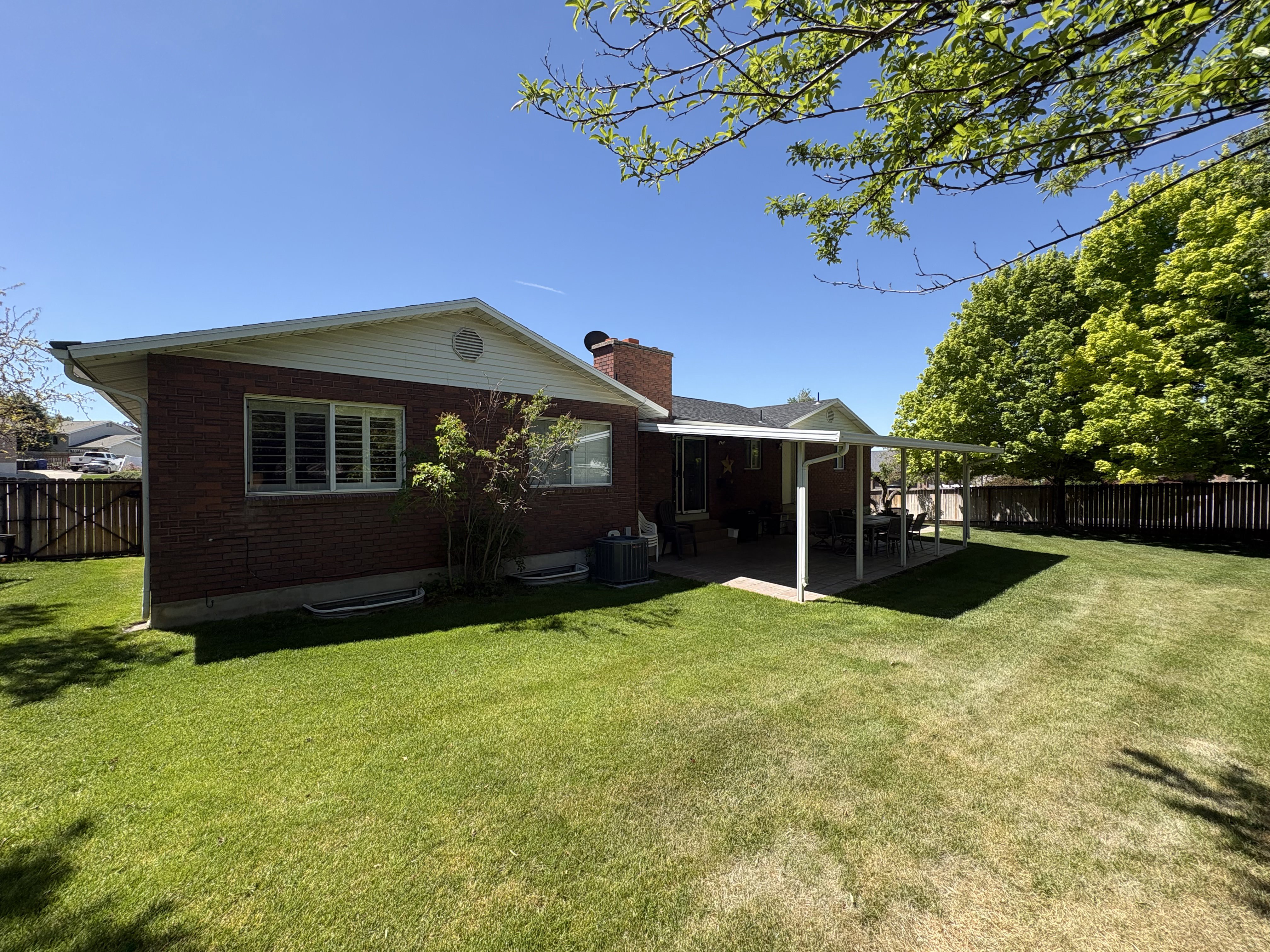 Spacious backyard featuring a covered patio, brick exterior, white vinyl siding, mature perimeter trees, and a wooden fence