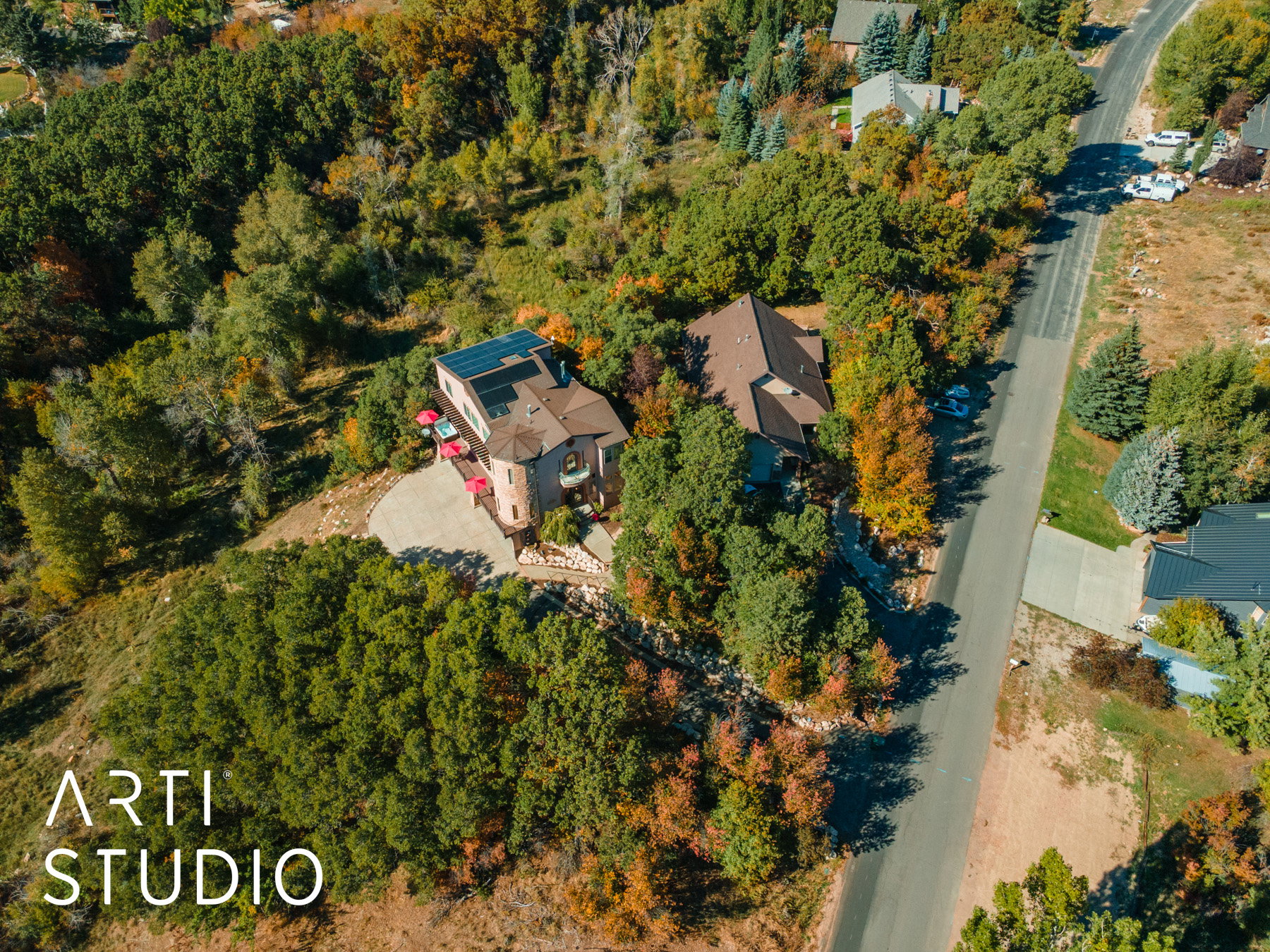 Hillside property featuring a stone turret exterior, a multi-paneled roof-mounted solar array, a curved paved driveway, and mature surrounding trees