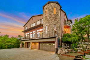 Stucco exterior featuring a stone-clad turret, multiple balconies with metal railings, a pergola-covered patio, and an integrated multi-car garage