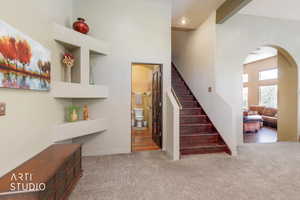 Carpeted entry area featuring a rich wood-finish staircase, built-in shelving, an arched doorway, and a visible powder room with wood-finish flooring
