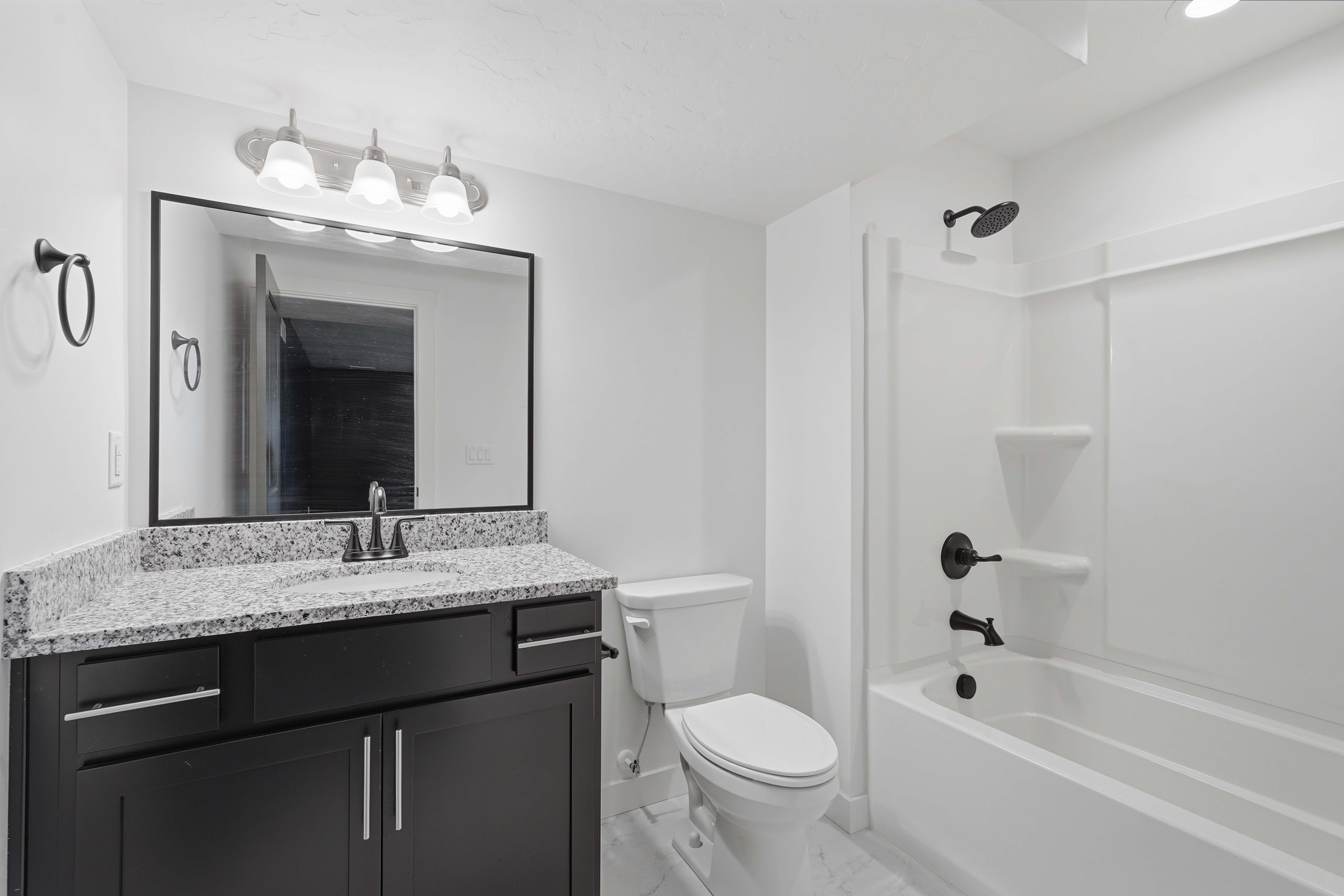 Full bathroom featuring a dark wood-finish vanity with a light speckled countertop, matte black fixtures, a framed mirror, and a full tub with shower