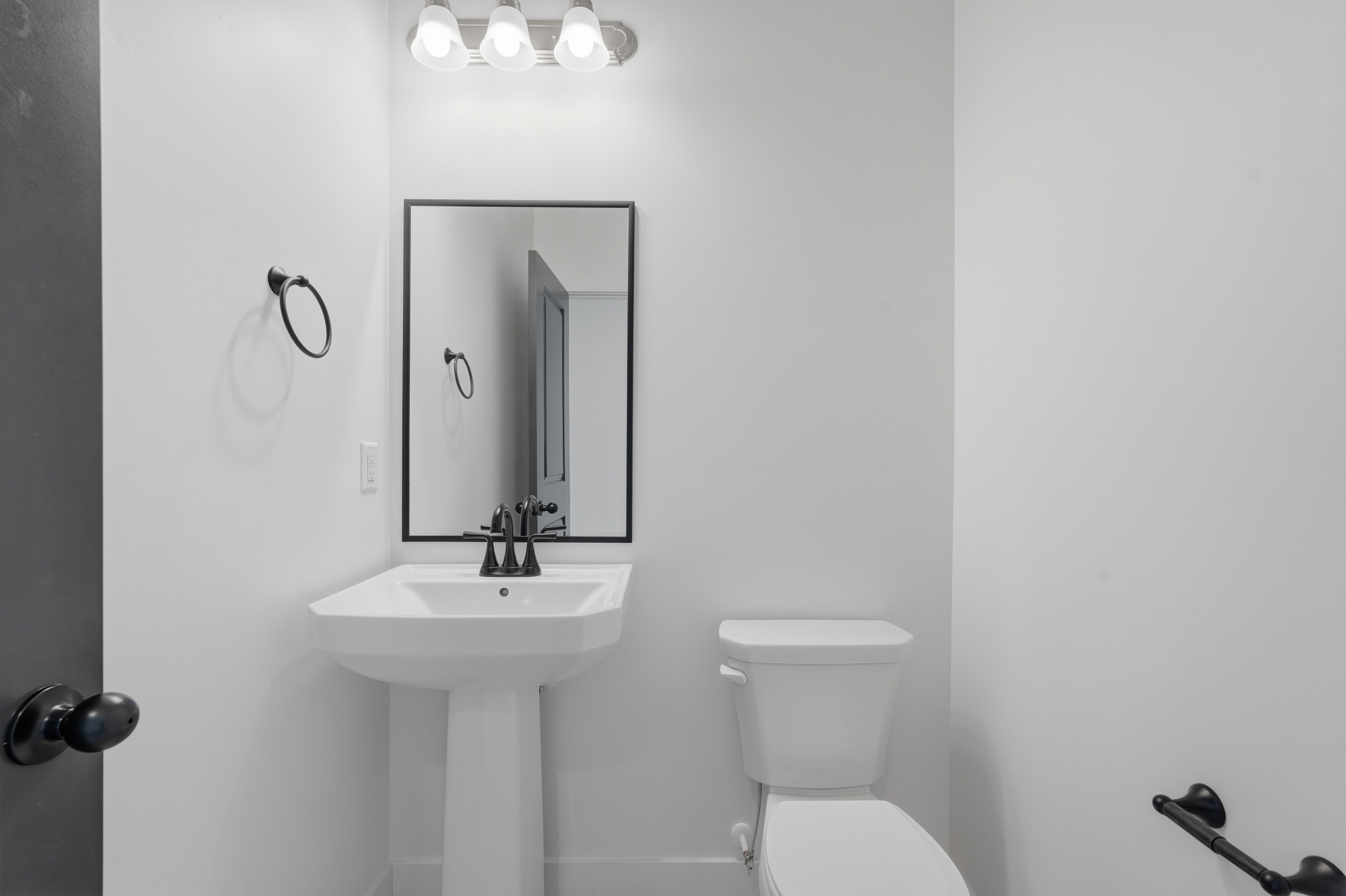Pedestal sink with matte black faucet and coordinating mirror, complemented by a white toilet and modern light fixture