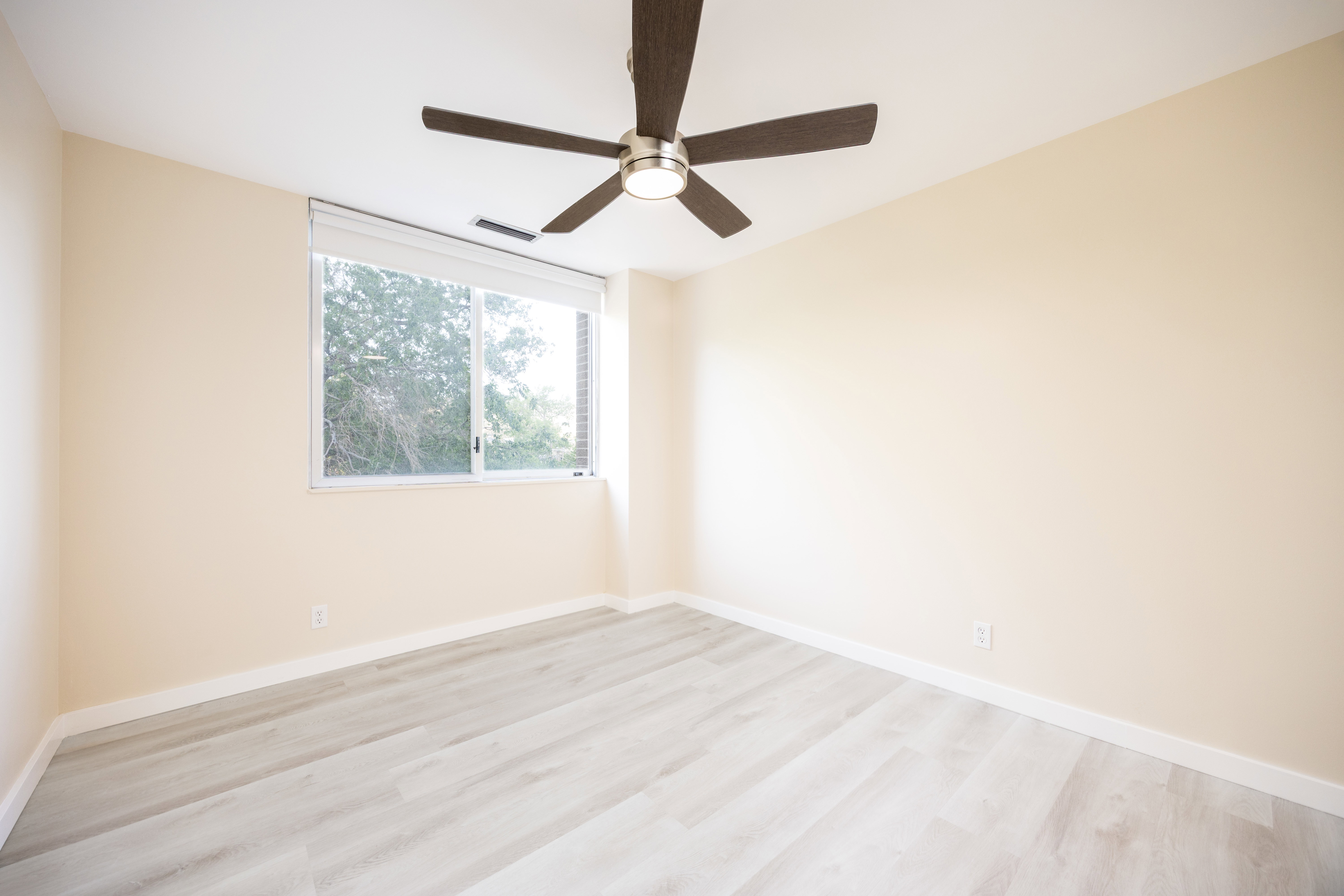 Bright room featuring a ceiling fan with integrated lighting, a large window with roller shades, light wood-finish flooring, and neutral wall paint