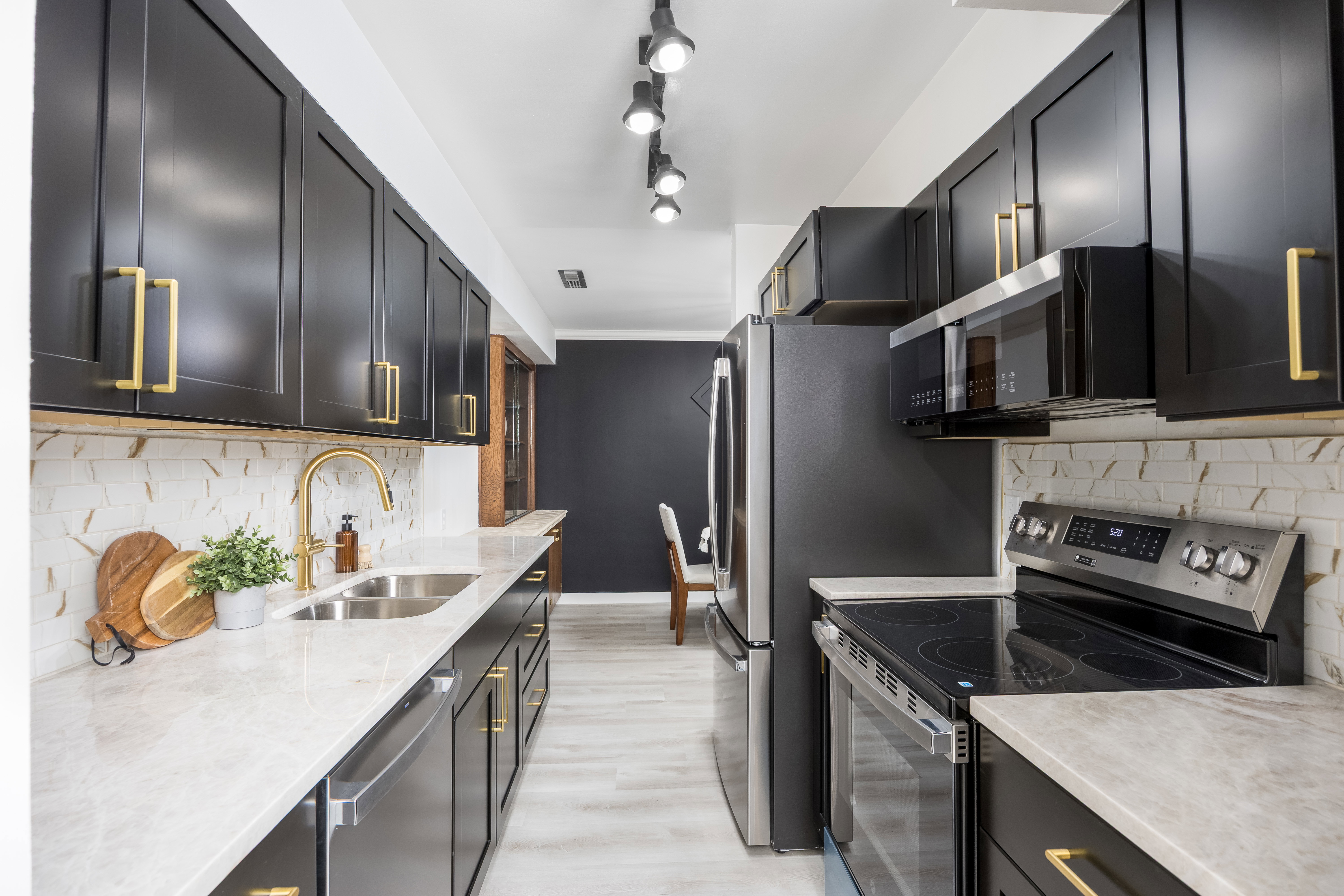 Kitchen featuring black cabinetry with gold-tone hardware, light-toned countertops, a stainless steel double-basin sink with a gold-tone faucet, a white and brown patterned backsplash, and light wood-finish flooring