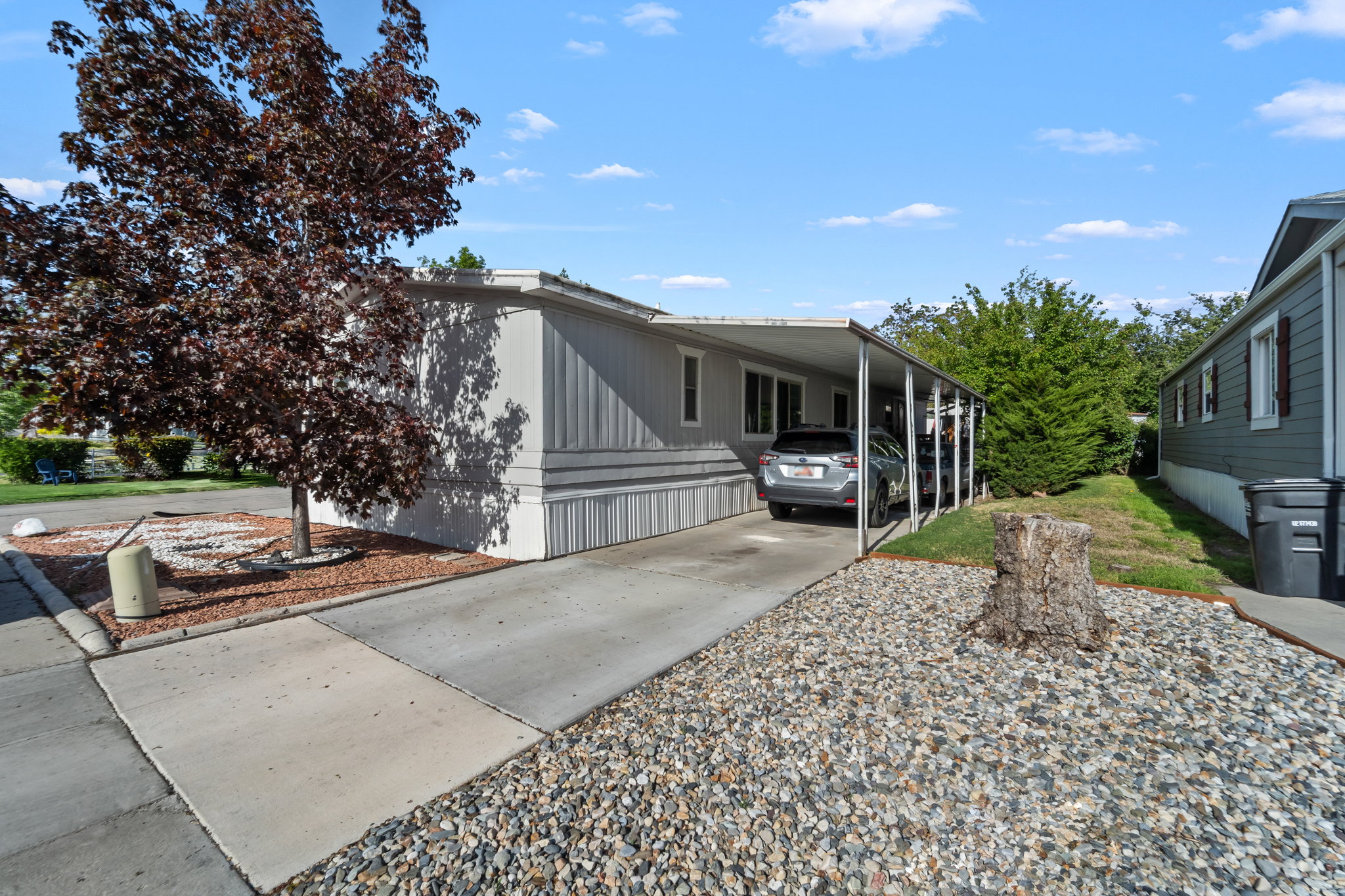 Manufactured home featuring light gray siding with white trim, an extended carport, a concrete driveway, a sidewalk, and low-maintenance rock landscaping