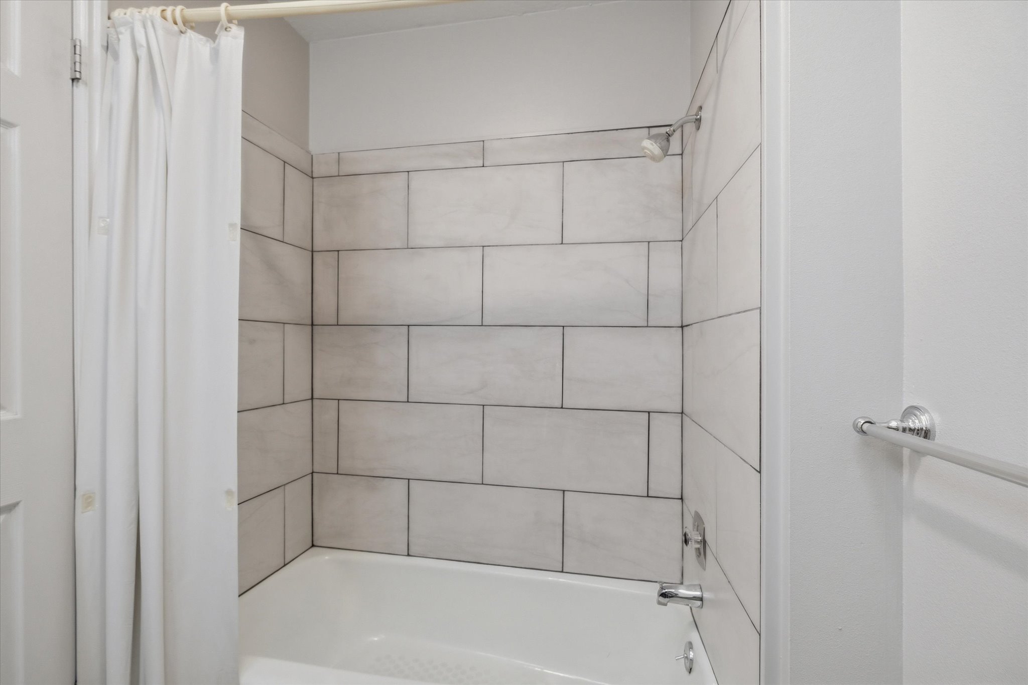 Full bathtub with light-toned rectangular tile surround, chrome showerhead, and matching chrome faucet