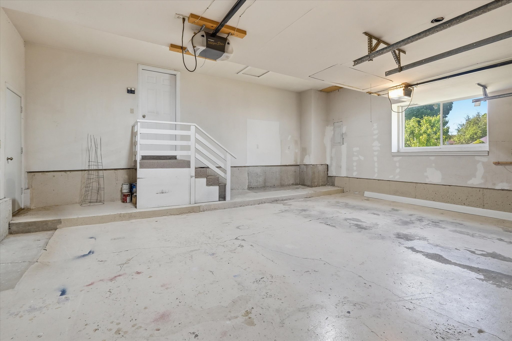 Spacious garage featuring a concrete floor, overhead garage door opener, interior access door, staircase, and a large window providing natural light. Storage above the garage doors.