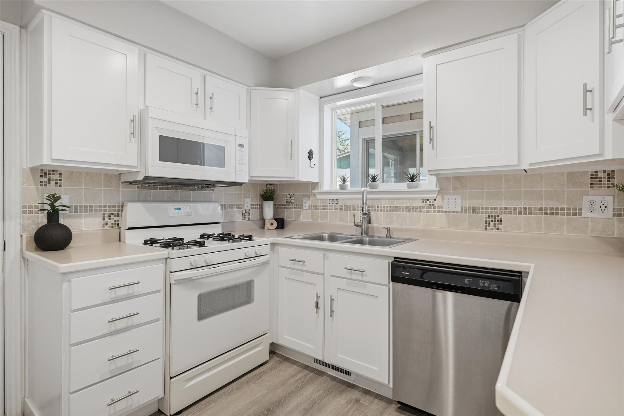 Kitchen featuring white cabinetry, light-toned countertops, tile backsplash with accent mosaic, stainless steel dishwasher, and a white gas range with overhead microwave