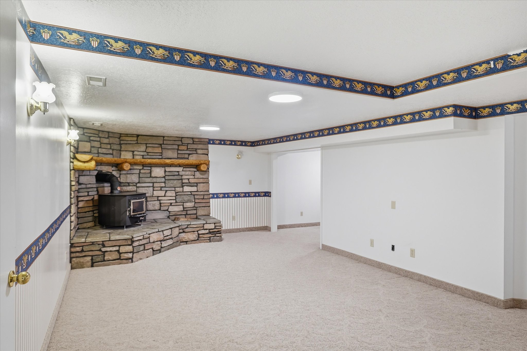 Stone fireplace with wood-burning stove, wall sconces, ceiling lighting, decorative wall border, and neutral carpet flooring