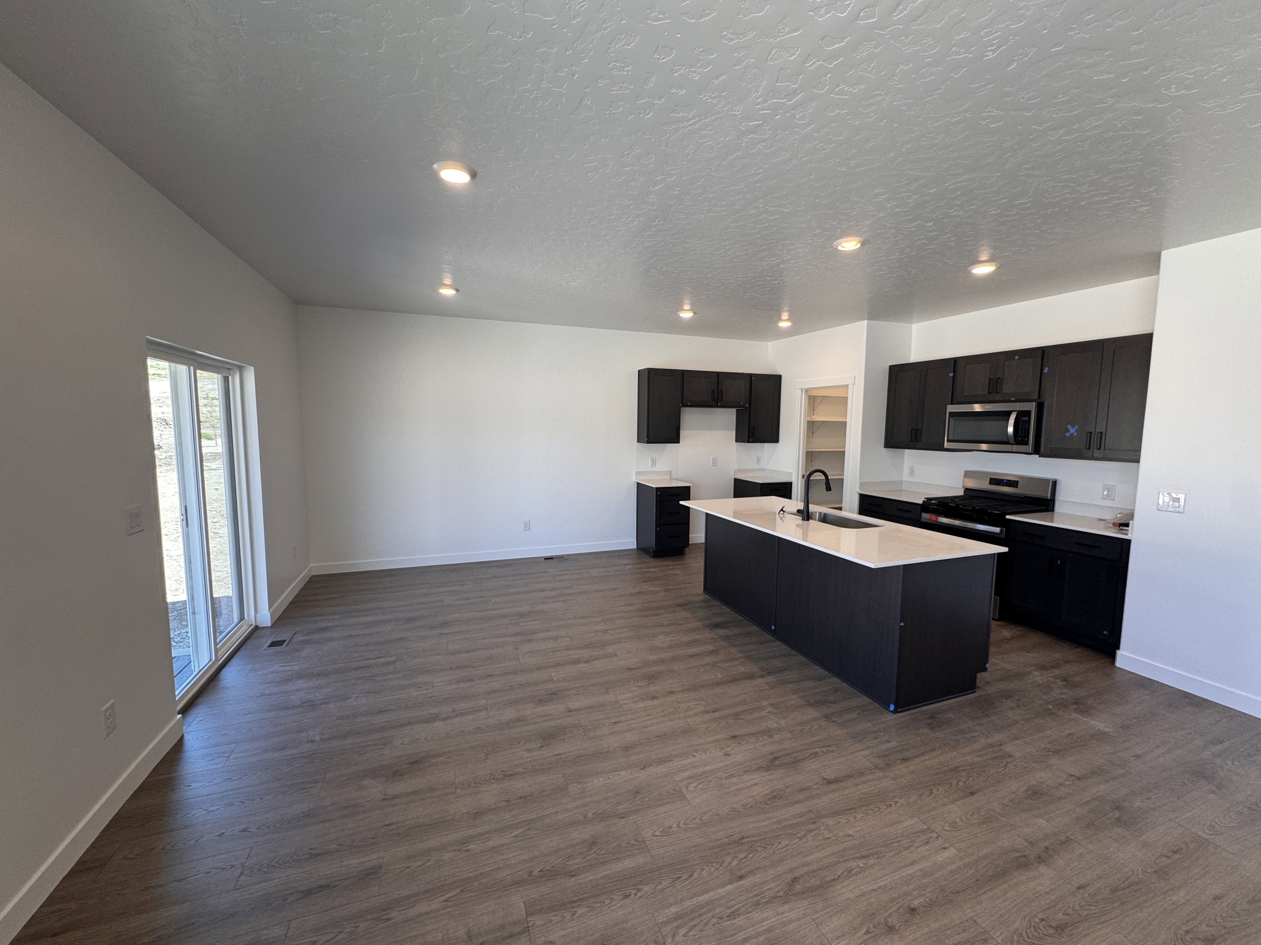 Open-concept kitchen and living area featuring dark cabinetry, a central island with an integrated sink, stainless steel appliances, and wood-finish flooring