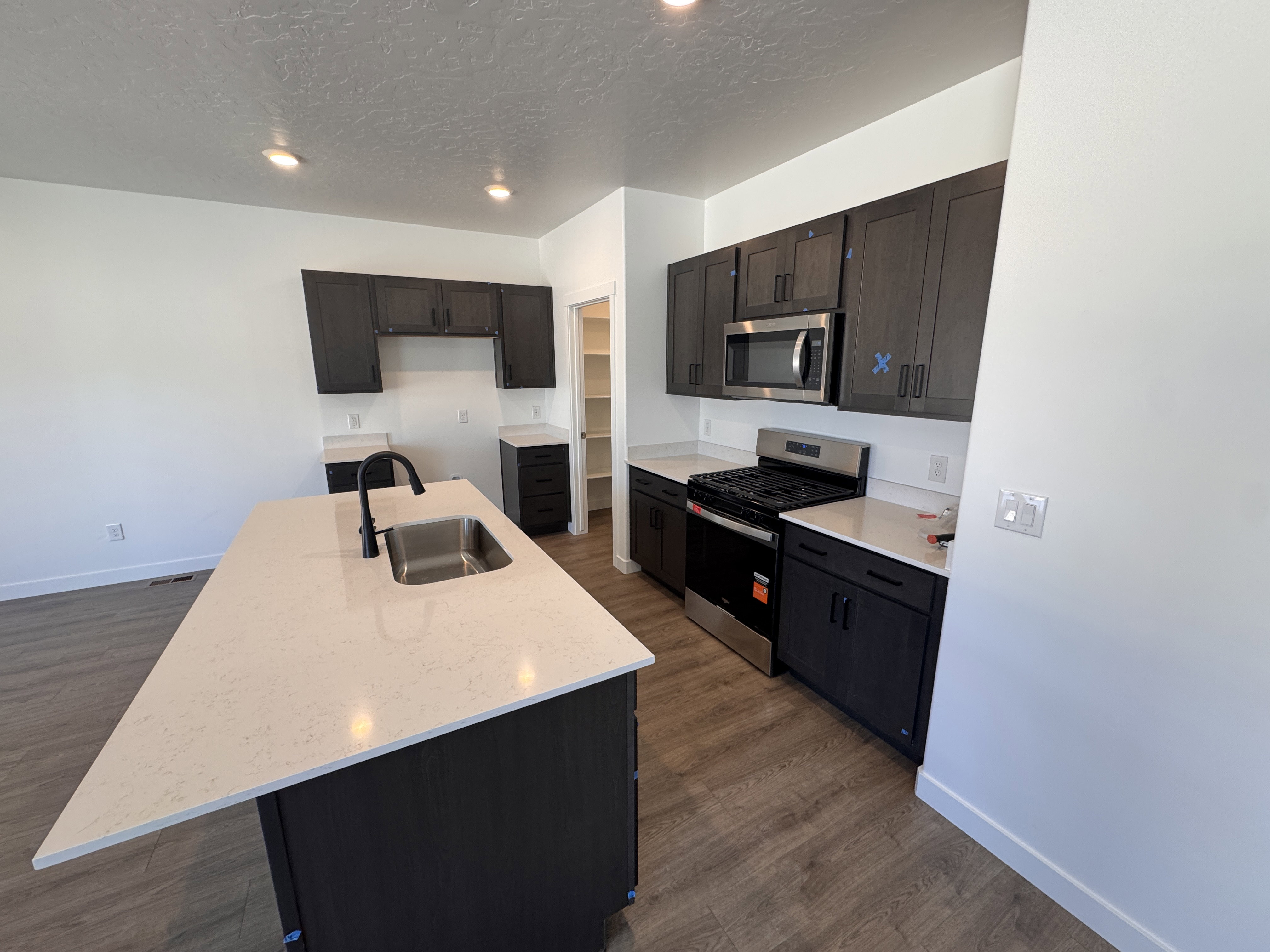 Kitchen featuring wood-finish flooring, dark wood-tone cabinetry, light-colored countertops, stainless steel appliances, and a central island with an undermount sink and matte black faucet